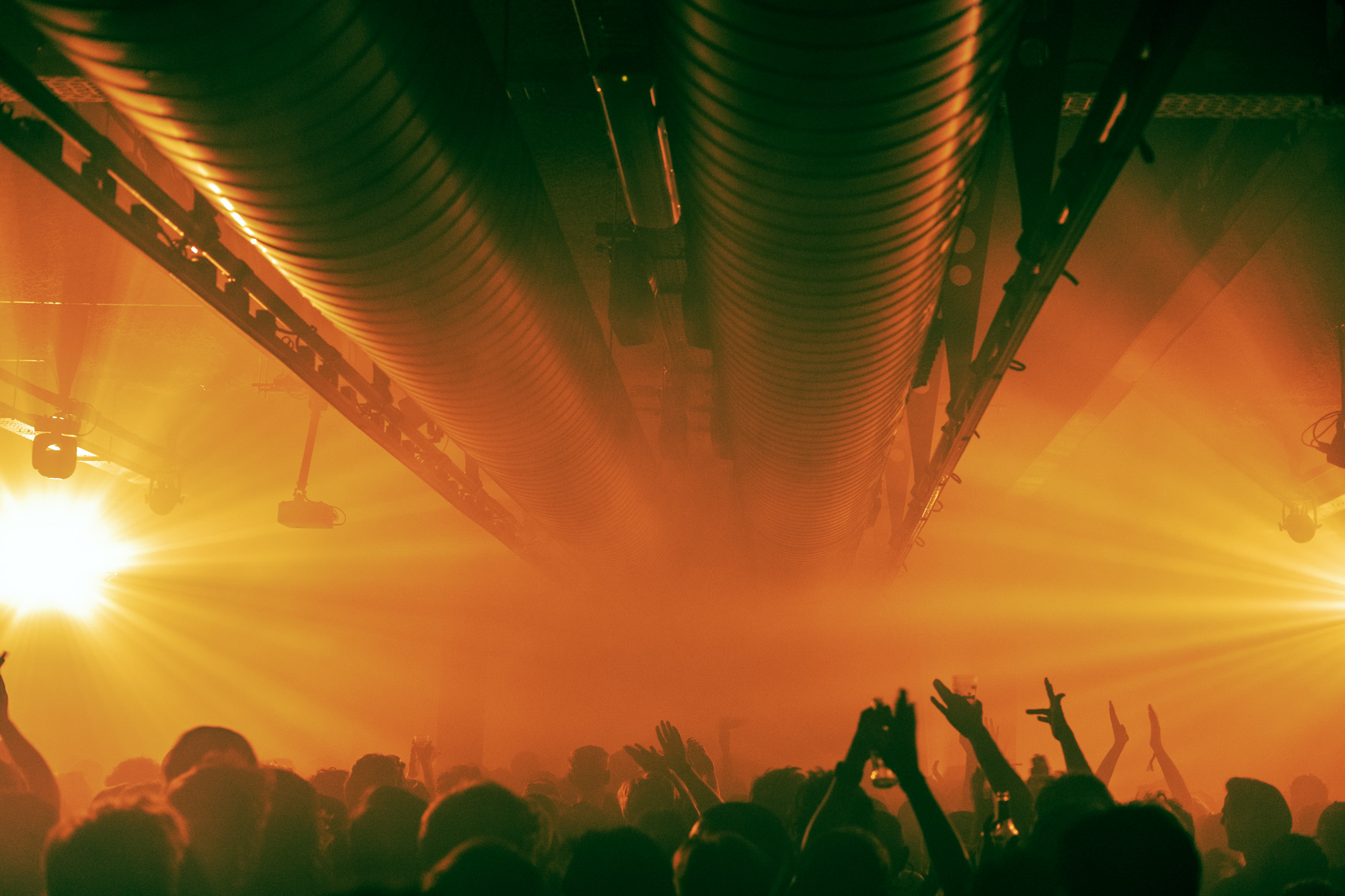 A photo of a well-attended club night. The space has an industrial look; you can see several pipes running along the ceiling. You can also see several visitors&rsquo; hands raised in the air.